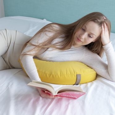 Młoda kobieta ubrana na biało w długich blond włosach leży na białej pościeli na w pozycji na brzuchu, wsparta na poduszce medytacyjnej w kształcie księżyca w kolorze tropikalno żółtym, czytając książkę.